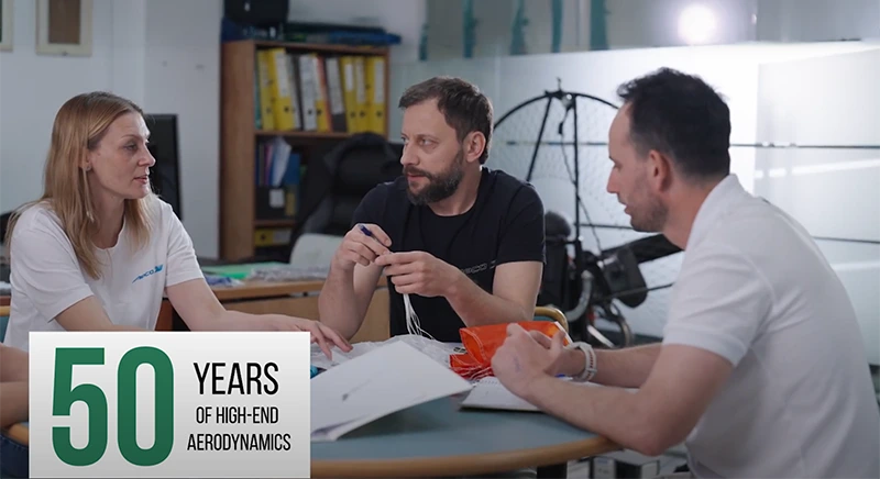 Tres personas del equipo técnico comentan alrededor de una mesa un pequeño velamen naranja, con el rótulo «50 Years of High-End Aerodynamics»