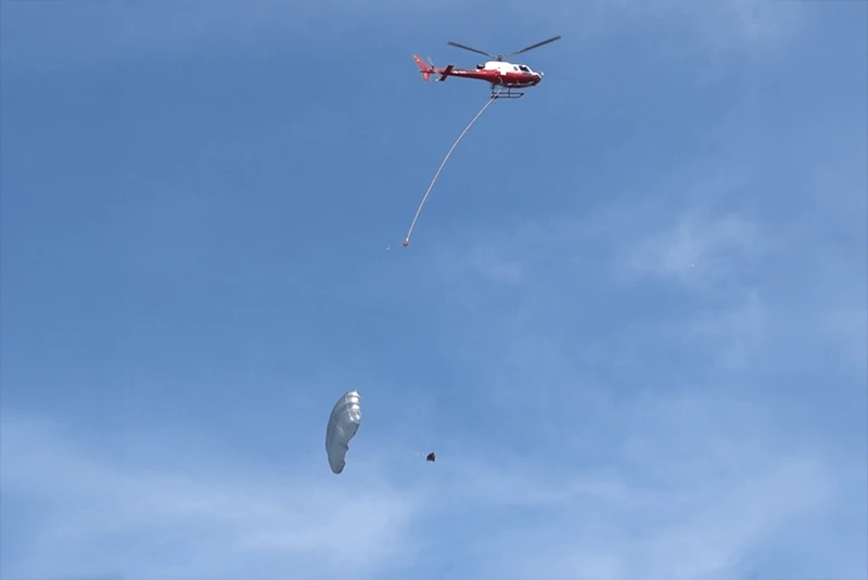 Elicottero in un cielo sereno che rilascia un carico su una fune lunga, con una velatura chiara già in apertura al di sotto