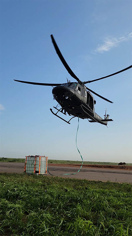 Helicóptero en vuelo estacionario sobre un aeródromo, con una cinta que baja hasta una carga en un contenedor de jaula en el suelo
