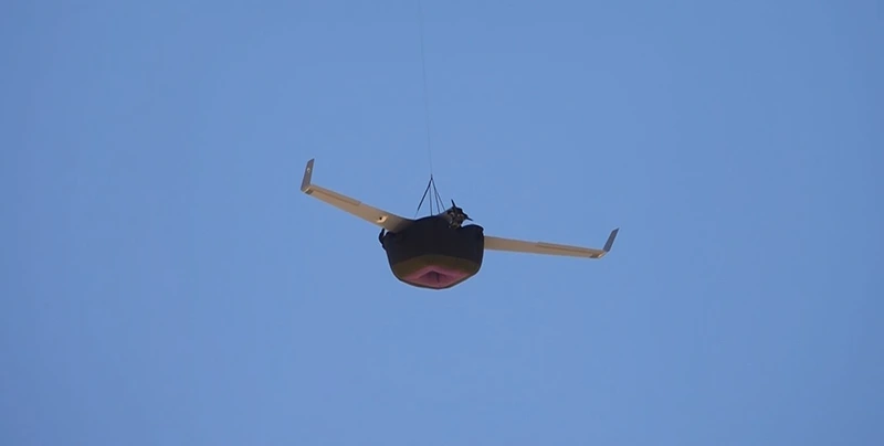 Dron de ala fija en vuelo con un paquete de recuperación oscuro bajo el fuselaje contra un cielo azul