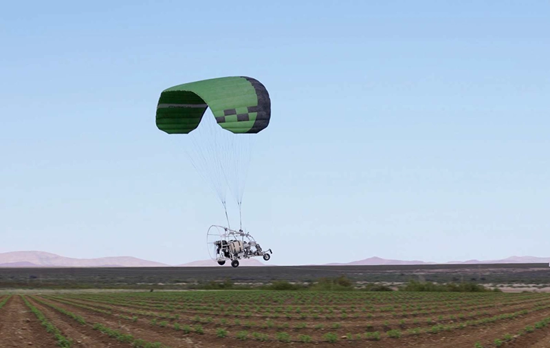 Trike no tripulado y sin asiento volando bajo una vela verde sobre cultivos en hileras, con colinas peladas en el horizonte