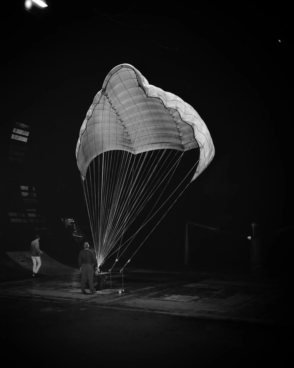 Fotografía en blanco y negro de un paracaídas Rogallo abierto en el suelo con dos personas debajo