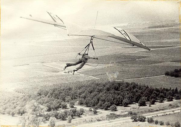 Fotografía antigua en blanco y negro de un piloto colgado boca abajo en un arnés bajo un ala delta, con campos y líneas de árboles muy abajo