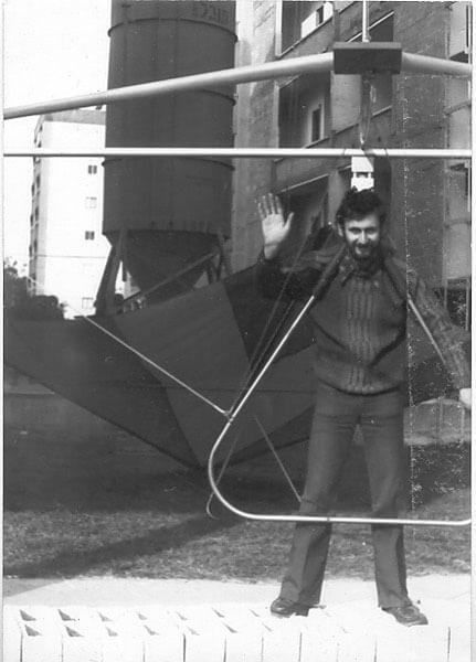 Fotografía en blanco y negro: un hombre saluda de pie dentro de la barra de control de un ala delta de vela oscura, delante de una nave industrial