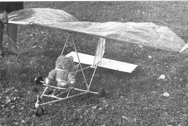 Fotografía en blanco y negro de un prototipo temprano de ala delta en un prado: estructura tubular sobre ruedas con vela de tejido y un peluche sentado en el asiento