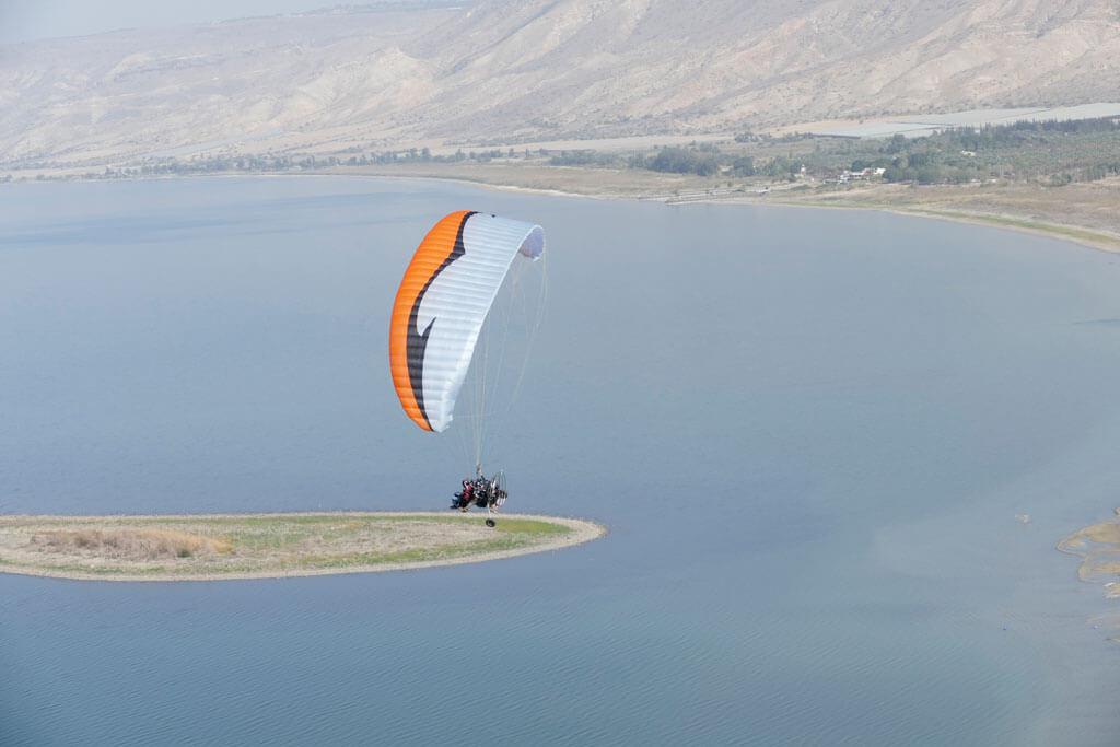 Trike volando bajo una vela naranja y blanca sobre un lago, con una lengua de tierra estrecha abajo y laderas peladas al fondo