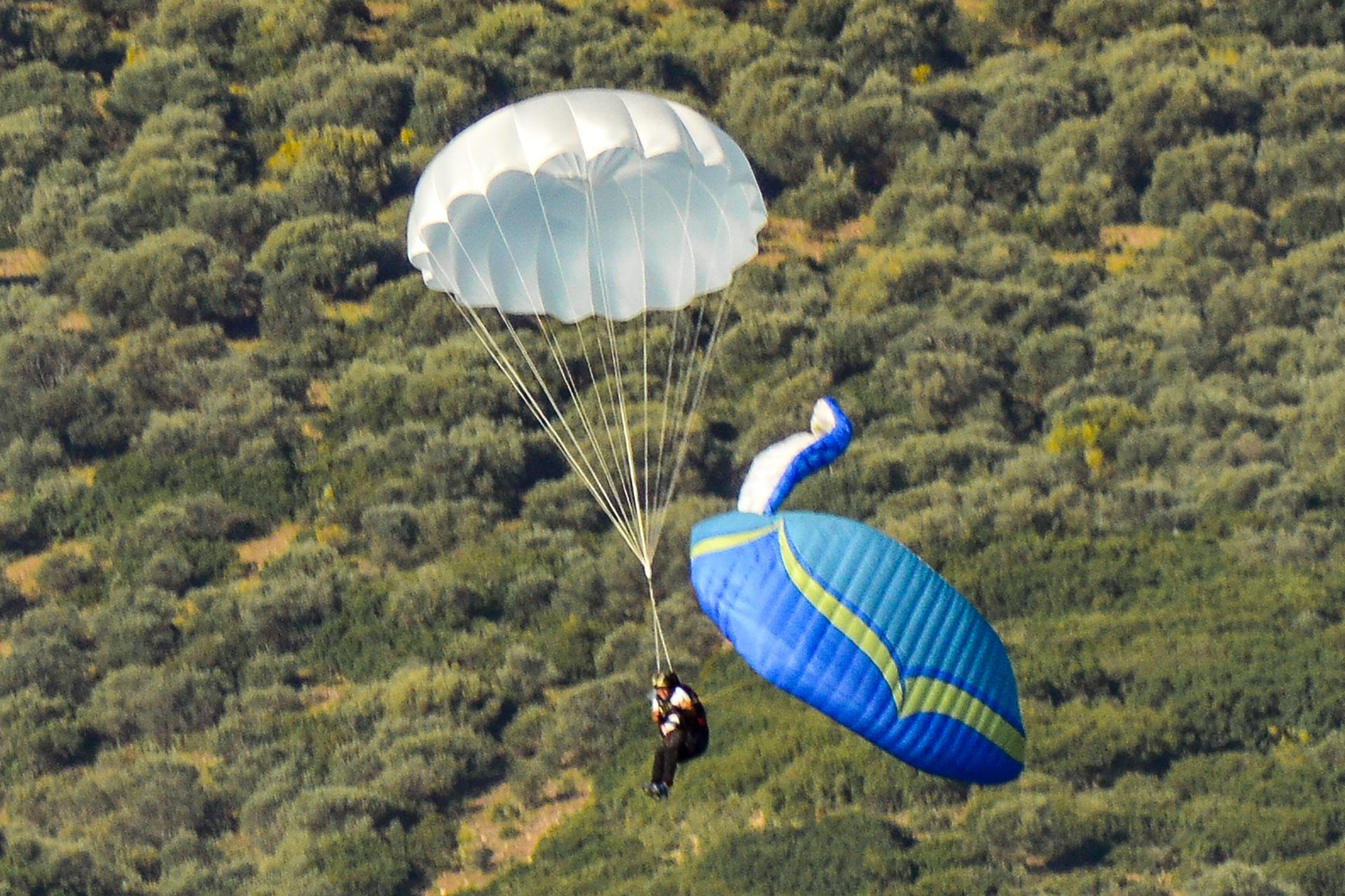 Campana de reserva blanca desplegada sobre un piloto, con el parapente azul plegado colgando al lado y terreno de matorral abajo