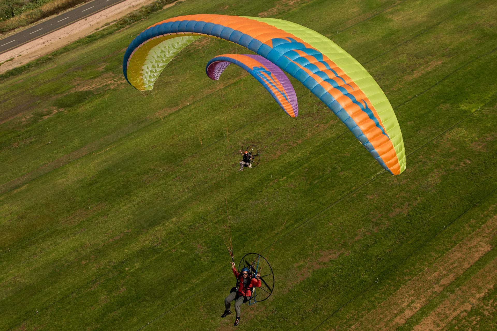 Vista cenital de tres velas de paramotor extendidas sobre una franja de hierba, con dos pilotos visibles en la imagen