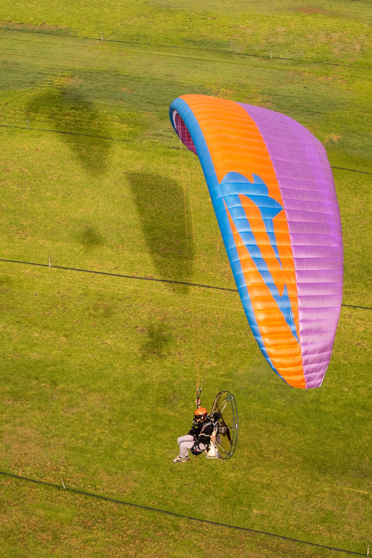Piloto con una unidad motriz a la espalda y jaula protectora volando bajo sobre una pradera, con la vela inclinada por encima