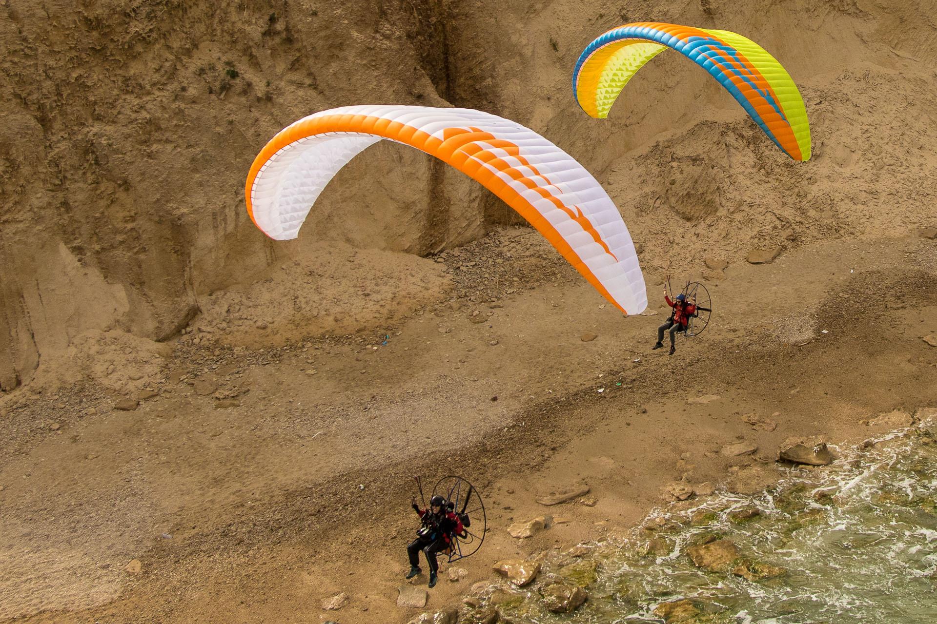Dos pilotos con unidades motrices a la espalda volando bajo junto a un acantilado de arena, con la rompiente abajo a la derecha