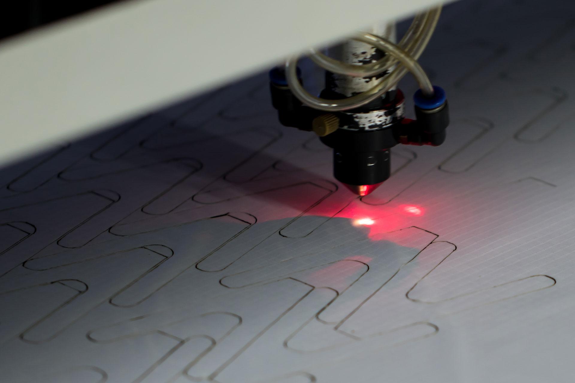 Cabezal láser cortando formas de panel en una pieza de tejido claro, con el haz brillando en rojo sobre la tela