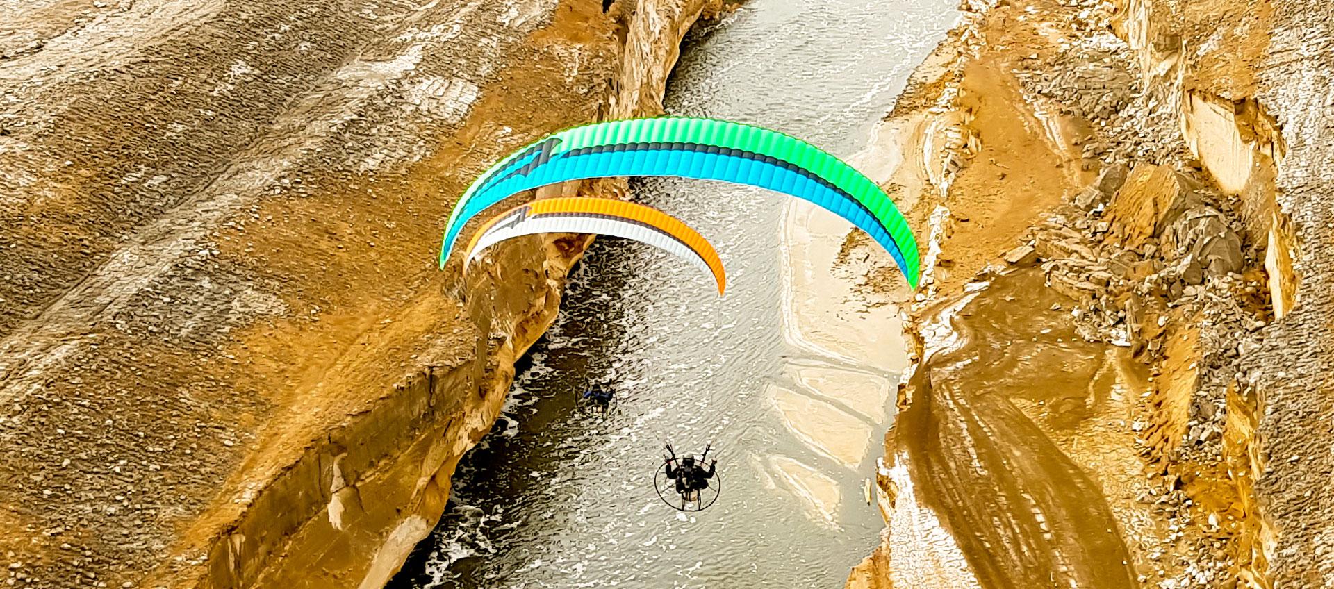 Dos pilotos de paramotor, uno bajo una vela verde turquesa y otro bajo una vela naranja, volando sobre un río entre paredes de roca clara