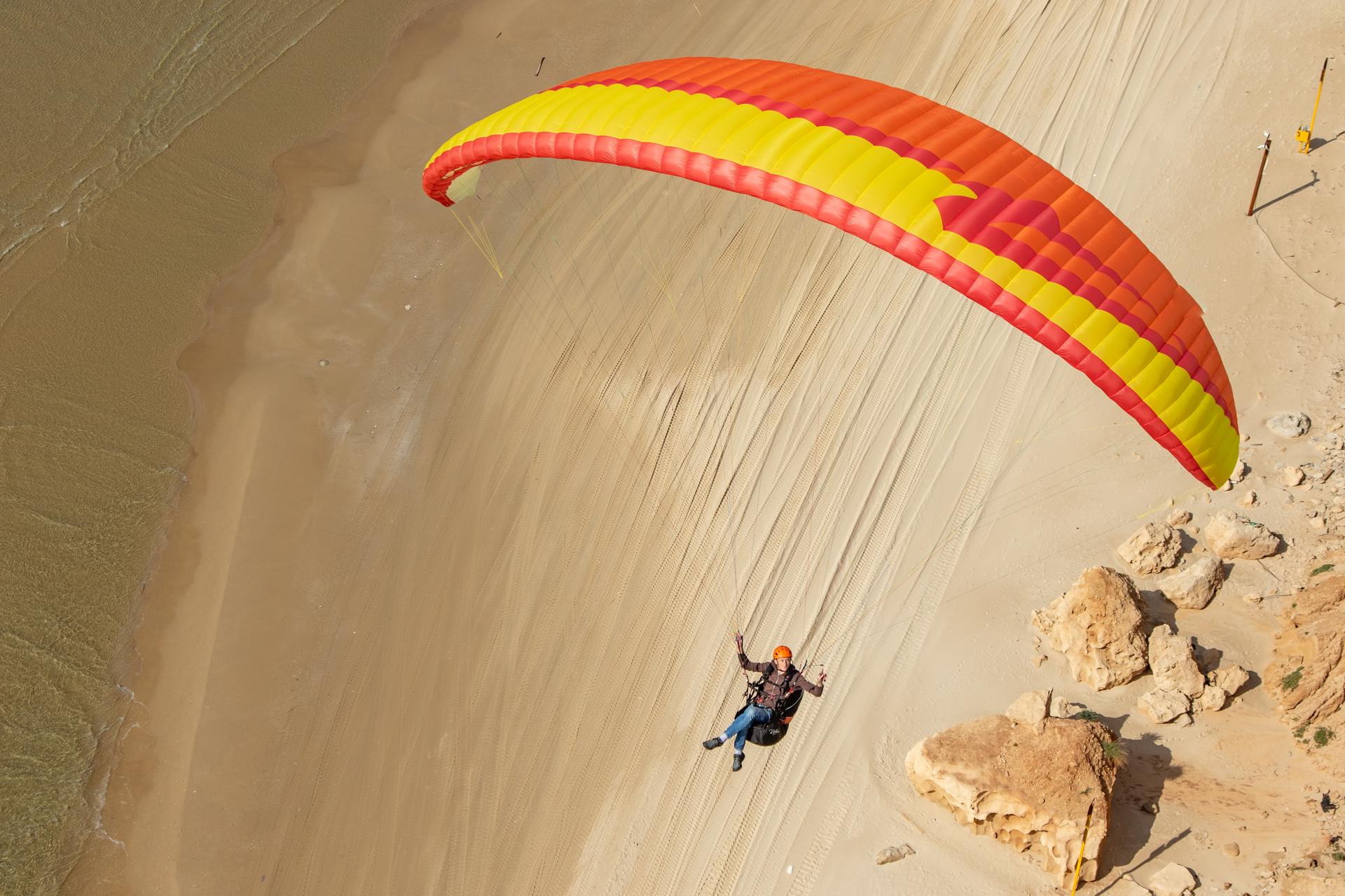 Piloto en un arnés bajo una vela de parapente roja y amarilla, volando bajo sobre una playa de arena a la orilla del agua