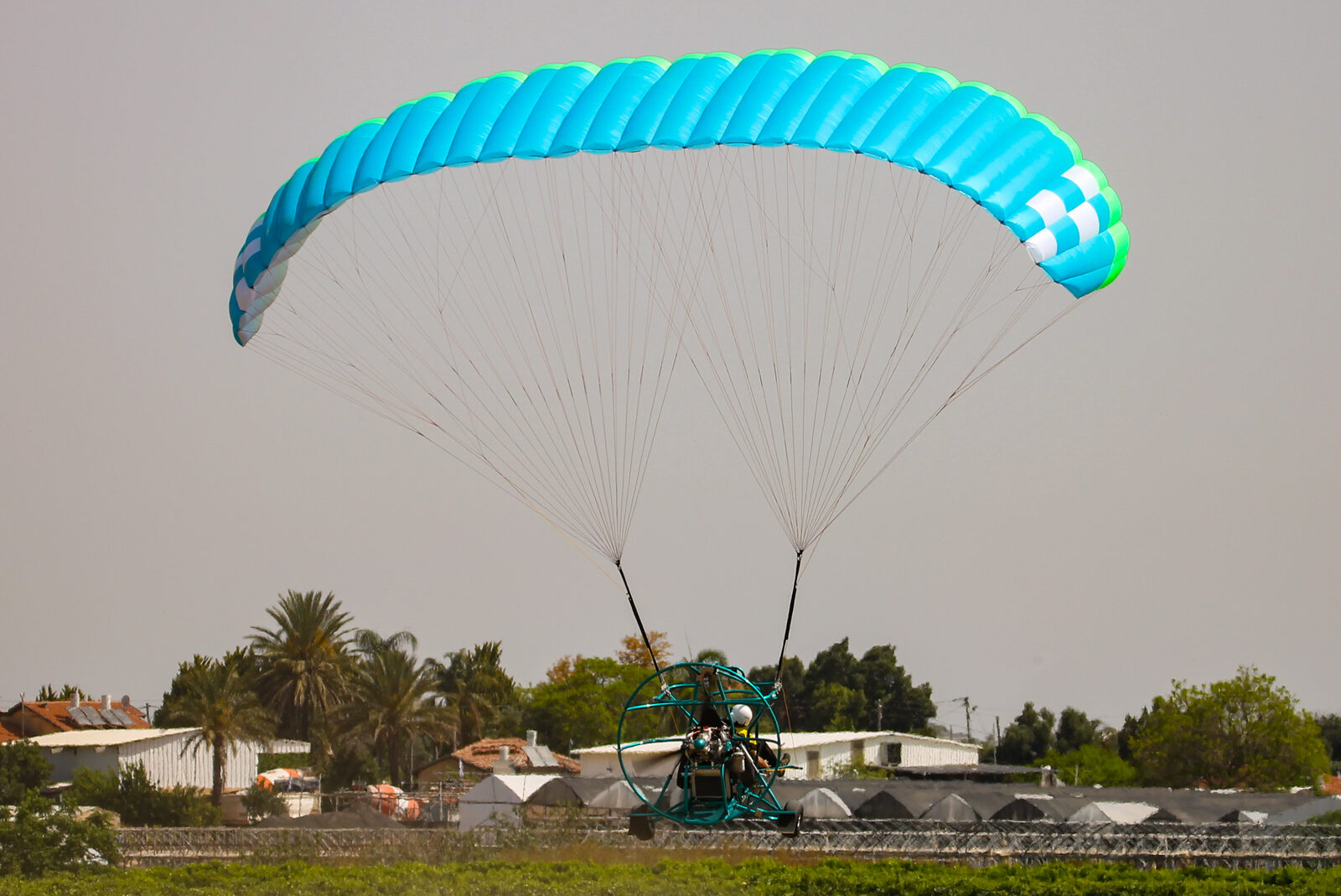 Trike volando bajo sobre campos, con palmeras, invernaderos y naves bajas al fondo