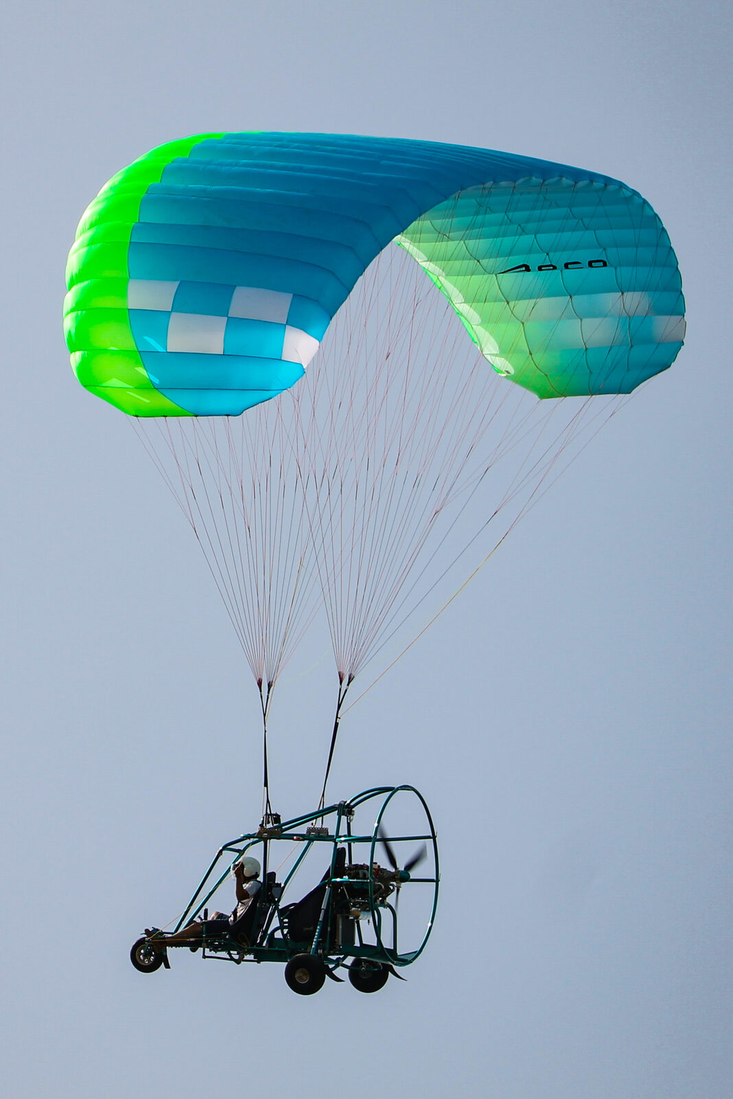 Aeronave de tres ruedas con estructura tubular, hélice propulsora enjaulada y un solo piloto, en vuelo bajo una vela verde y azul
