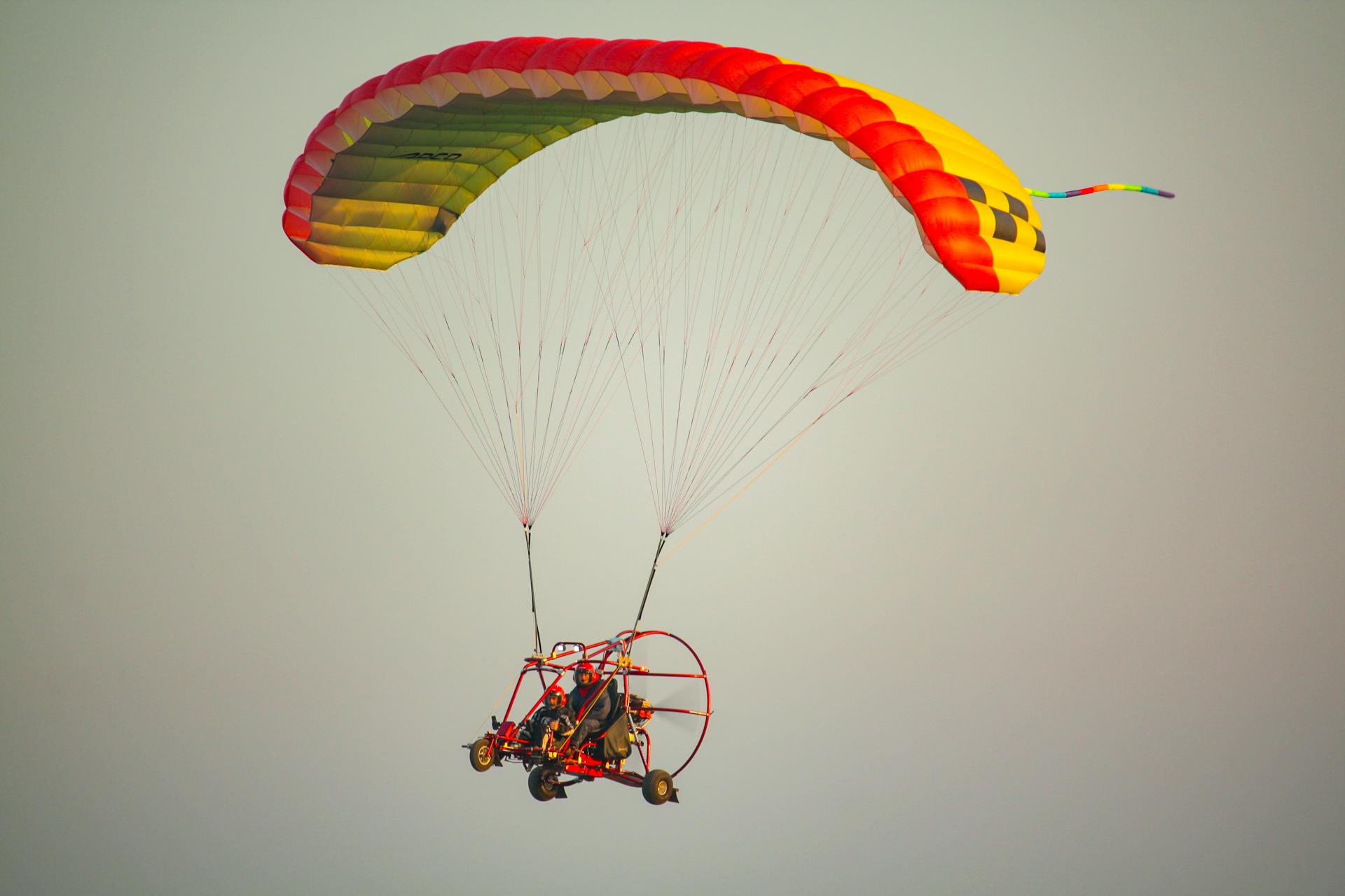 Trike de estructura roja con dos ocupantes en asientos en tándem, en vuelo bajo una vela roja y amarilla
