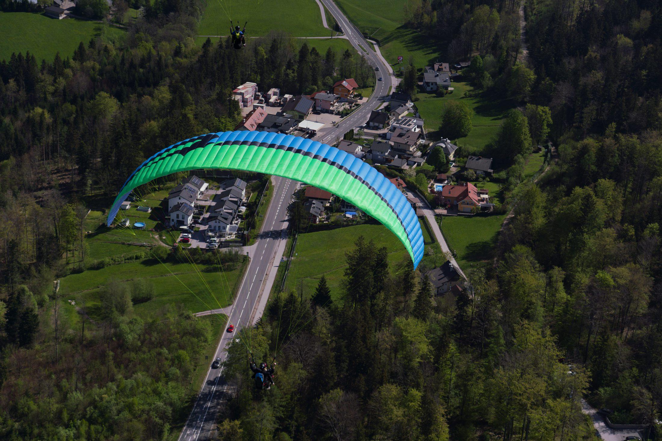 Vela de parapente azul y verde sobre los prados, bosques y casas de un valle, con un segundo piloto más alto por detrás