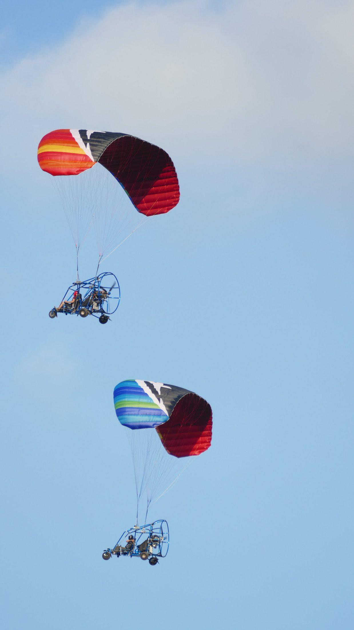 Dos trikes de paramotor, uno bajo una vela roja y amarilla y otro bajo una vela azul y verde, volando uno tras otro en un cielo azul claro