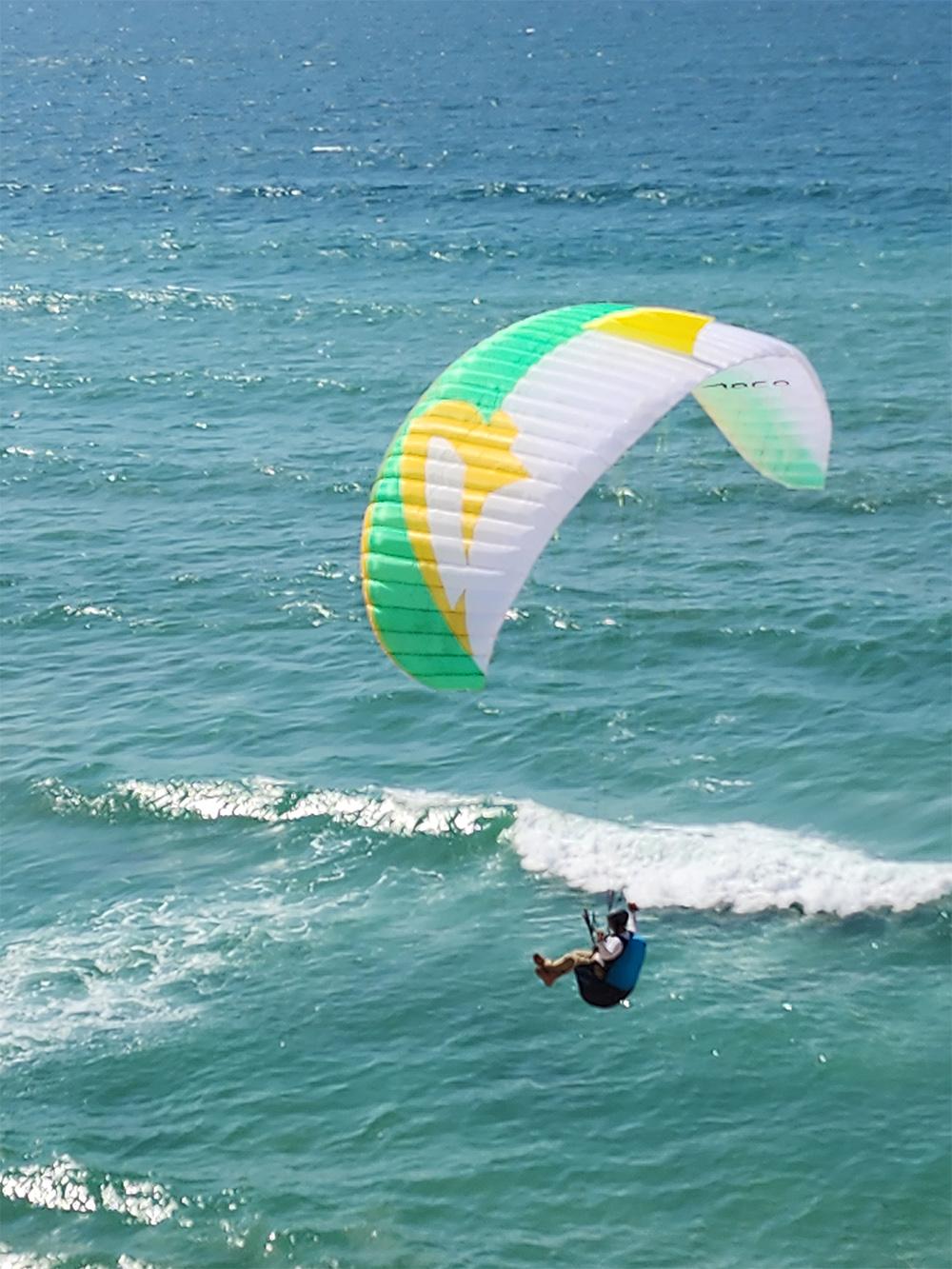 Piloto bajo una vela verde, amarilla y blanca sobre la rompiente, con una ola rompiendo debajo